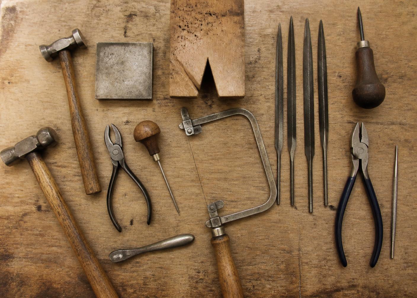 Worn jeweller's hand tools laid out on a scarred wooden bench: hammers, files, saw frame and pliers
